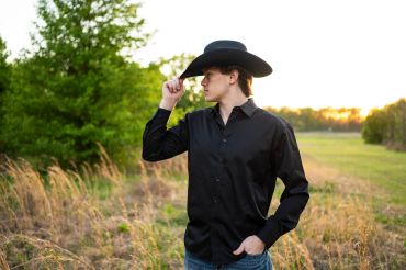 Young man in a black shirt and cowboy hat standing in a field at sunset.