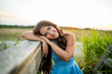 A woman in a blue dress leaning on a wooden railing outdoors at sunset.