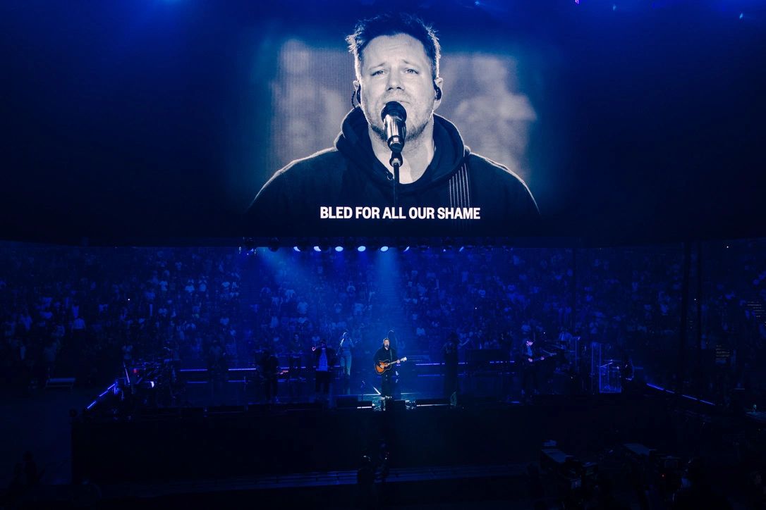 A musician performs on stage with a large screen behind showing his face and lyrics.