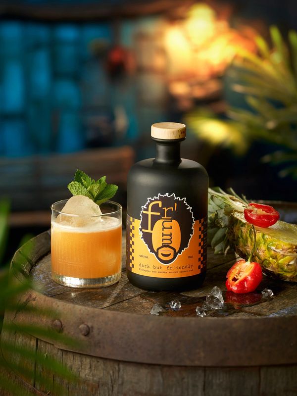A cocktail next to a bottle of Fr’um with pineapple and chilli on an old barrel in a Tiki bar.