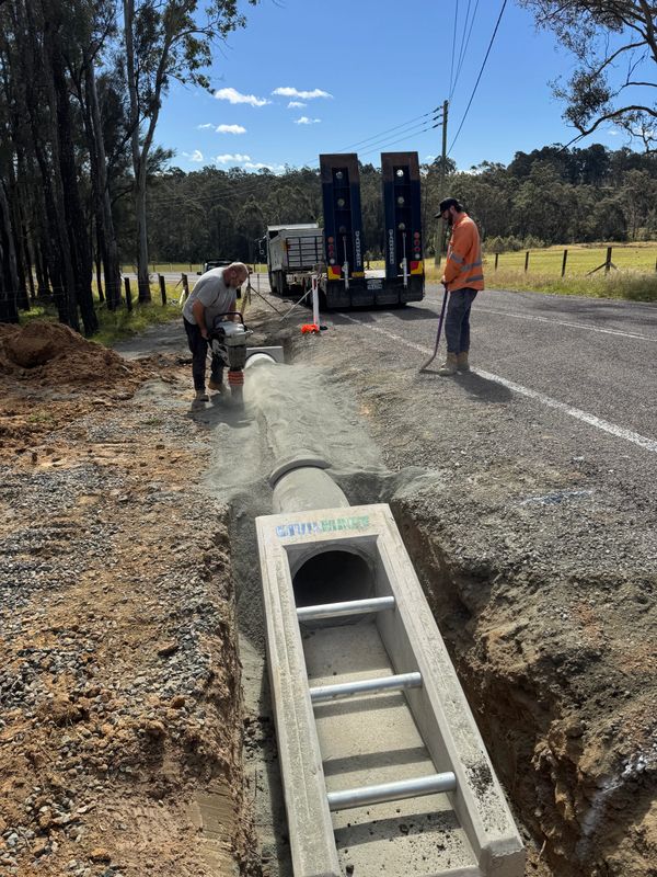 Two workers installing a concrete drainage pipe by the roadside under a clear sky.