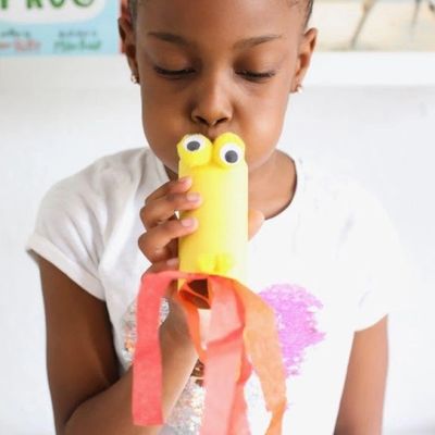 Child blowing into a yellow craft with googly eyes and red streamers.