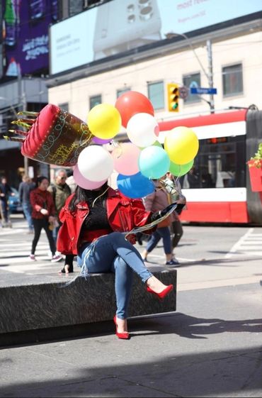 Person in red jacket holding colorful birthday balloons, sitting on a bench in city.