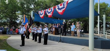 A patriotic ceremony with a color guard and audience under red, white, and blue bunting.