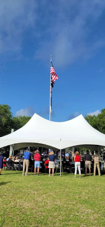 People gathered under a white tent with flags on a sunny day.