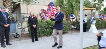 A man in sunglasses and a blazer stands before patriotic wreaths during a memorial event.