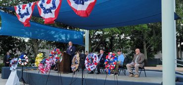 A man speaks at a memorial event with patriotic decorations and floral wreaths.