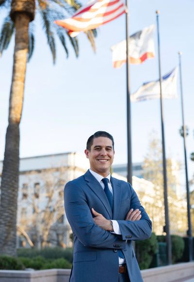 Confident man in a blue suit stands outdoors with flags and palm trees in the background.