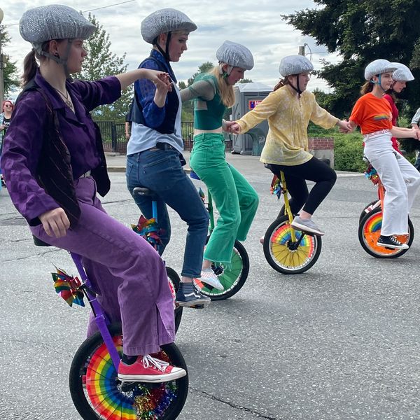 A group of six people wearing shiny helmets riding unicycles decorated with rainbow patterns, holding hands in a line.