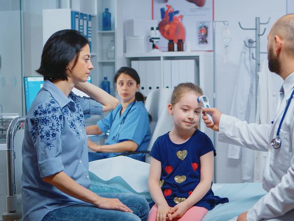 Doctor checking a young girl's temperature in a clinic while her mother watches.