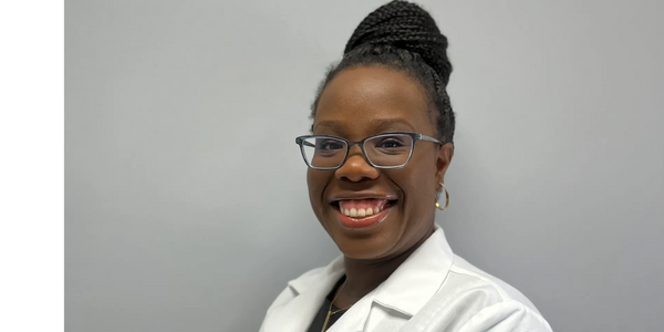 Smiling woman wearing glasses and a white lab coat against a gray background.