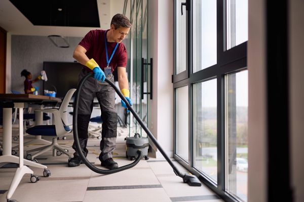 A janitor vacuuming the floor near large office windows.