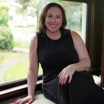 A smiling woman in a black dress sitting by a window.