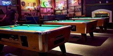 Dimly lit pool hall with three pool tables and neon signs.