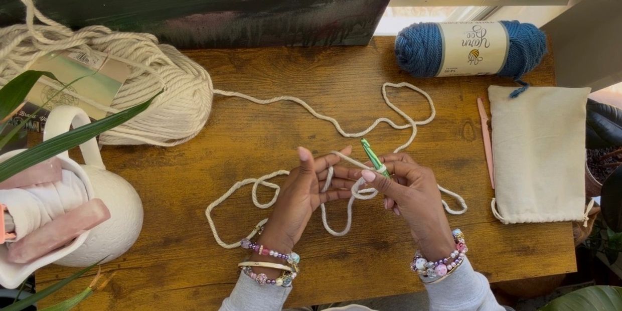 Person crocheting with yarn on a wooden table surrounded by plants and crafting tools.