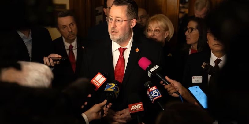 Man in suit and red tie speaks to reporters with microphones in a press conference.
