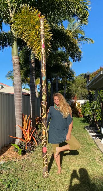 Woman posing barefoot in garden with decorated didgeridoo and palm trees.