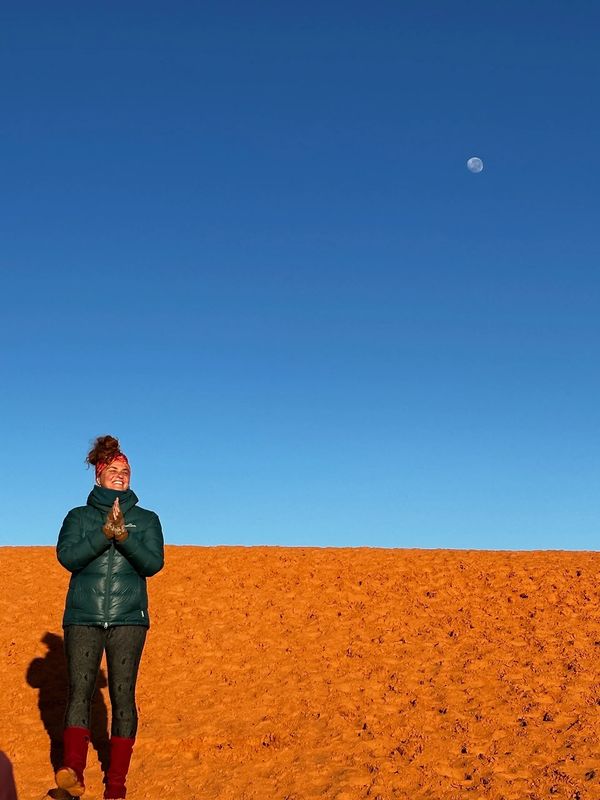 A person in a green jacket stands on red sand under a clear blue sky with the moon visible.