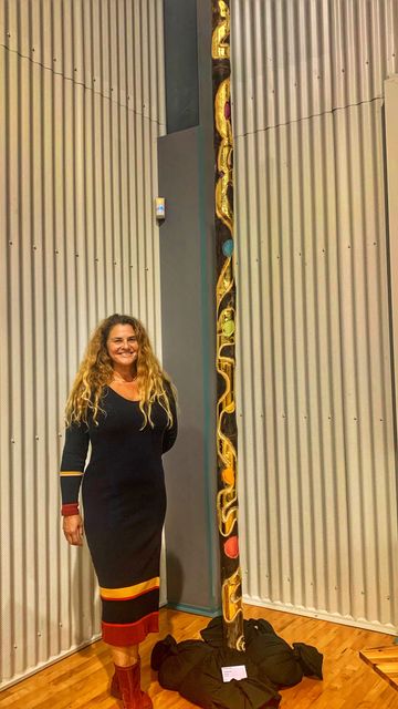 A woman stands smiling next to a tall decorated totem pole indoors.