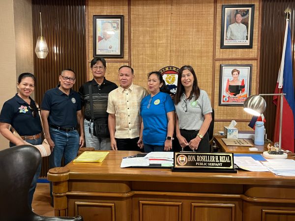 Group of six individuals standing behind a public servant's desk with nameplate Hon. Roseller H. Rizal.
