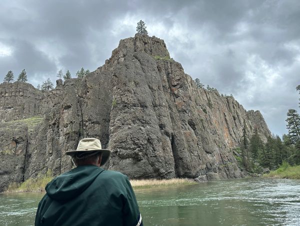 Person in a hat gazing at a towering rocky cliff by the river under a cloudy sky.