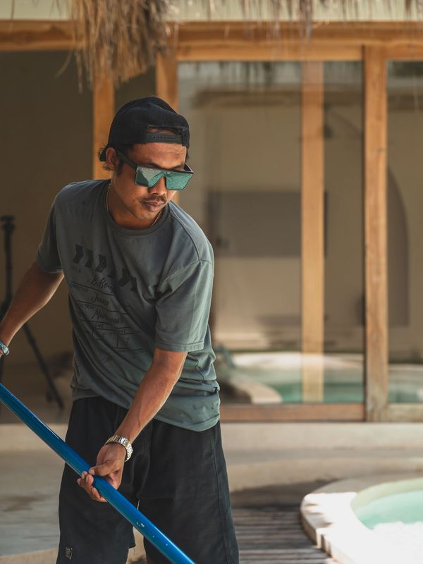 Man cleaning a pool with a net pole, wearing sunglasses and a cap.