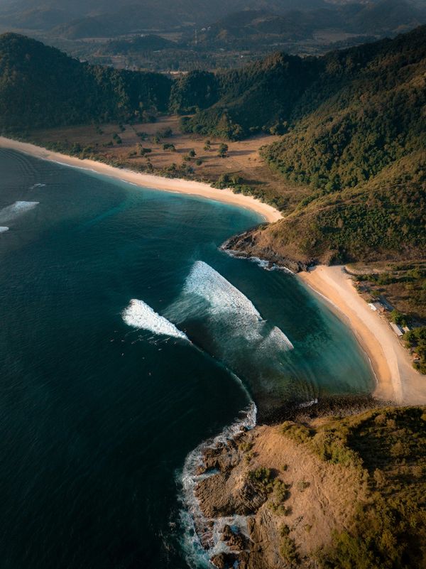 Aerial view of a coastal bay with waves, sandy beaches, and forested hills.