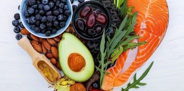 Heart-shaped arrangement of healthy foods like salmon, avocado, berries, nuts, and greens.