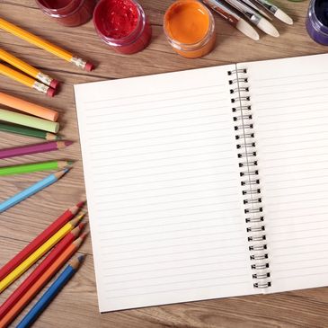 Open spiral notebook with colored pencils and paint jars on wooden table.