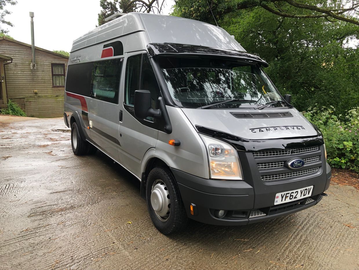 Silver Ford campervan parked on a wet driveway beside trees and a wooden building.
