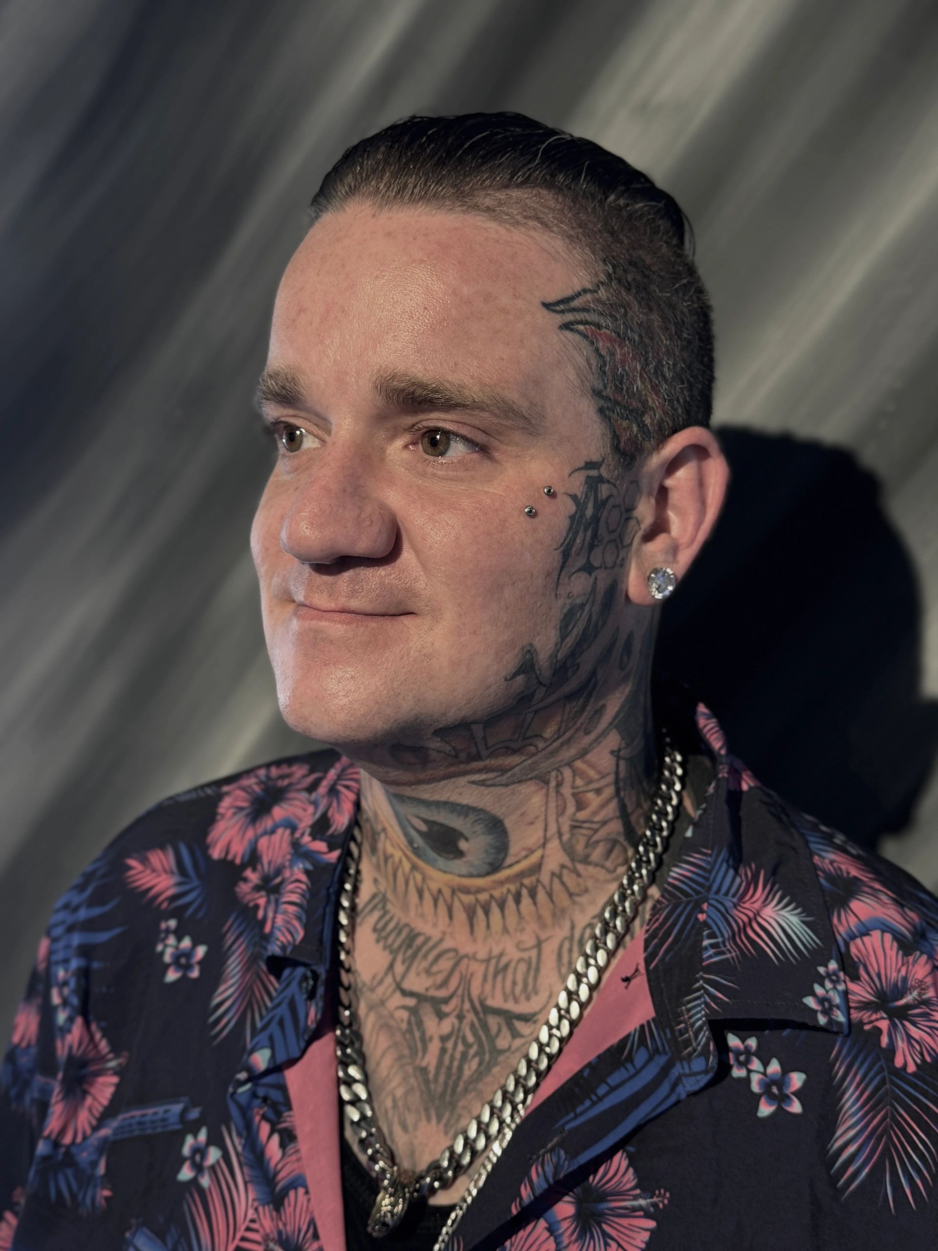 Man with facial tattoos and piercings wearing floral shirt and chains.