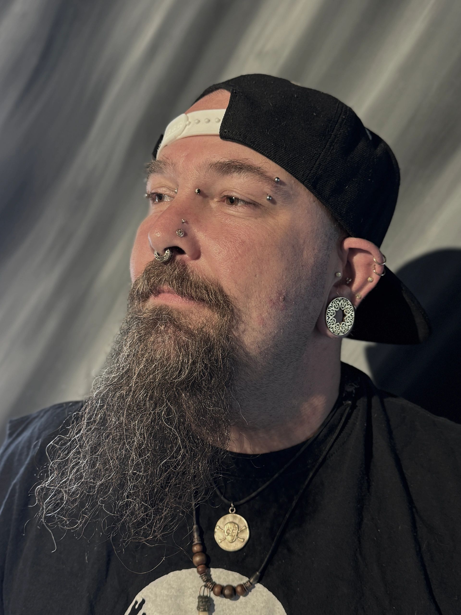 Man with multiple facial piercings and a beard wearing a cap.