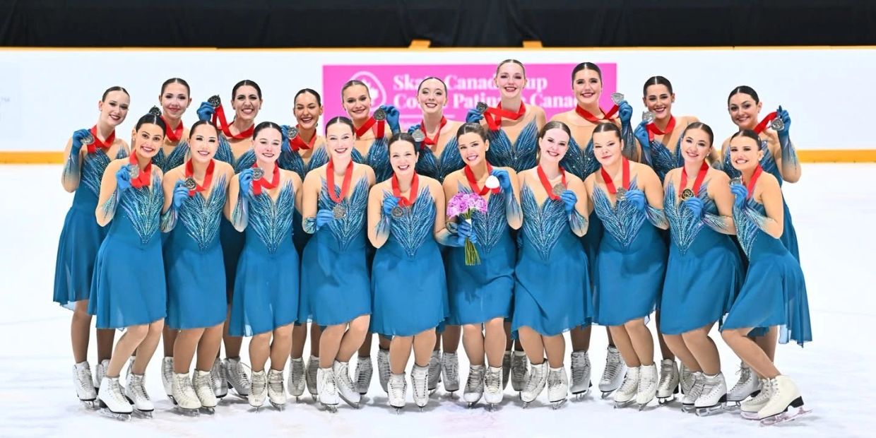 Synchronized skating team proudly displays medals in matching blue dresses on ice rink.