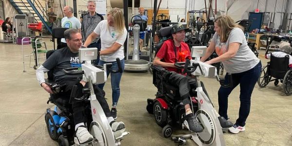 Two men in wheelchairs exercise with assistance in a gym.