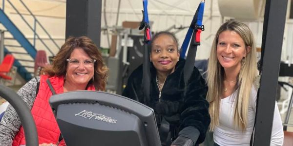 Three women smiling at a gym with exercise equipment.