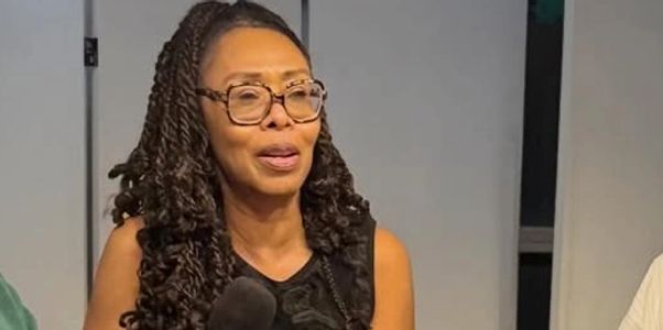 Woman with long braided hair and glasses speaking into a microphone.