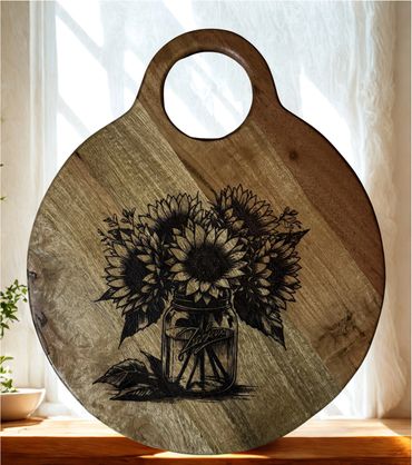 Round wooden cutting board with a sunflower bouquet engraving.