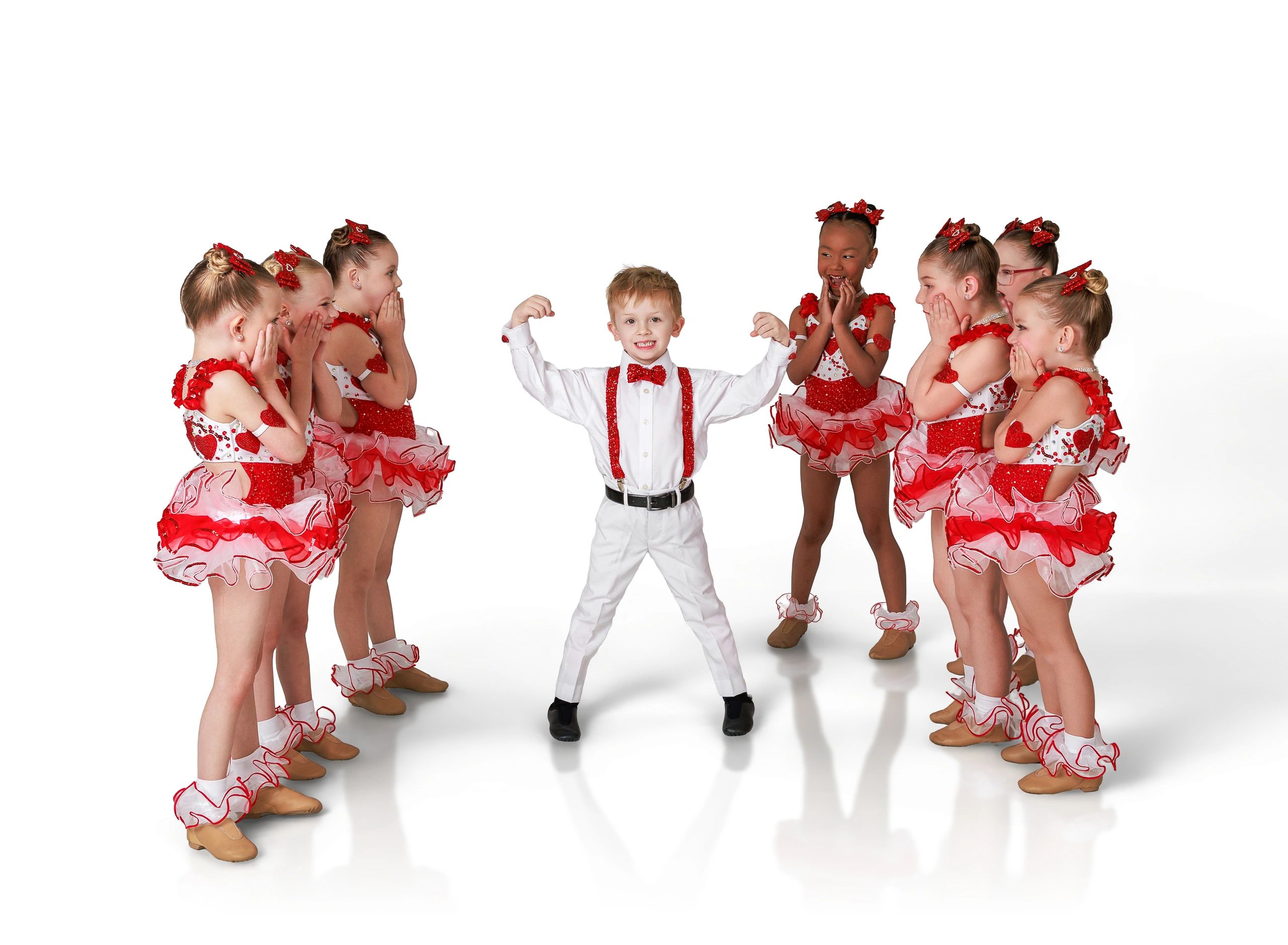Young boy flexes muscles surrounded by amazed girls in red dance costumes.