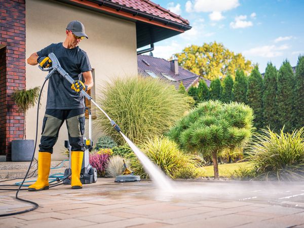 A person uses a pressure washer on a stone patio surrounded by green shrubs