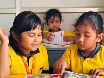 Three schoolgirls in yellow uniforms reading books at a desk.
