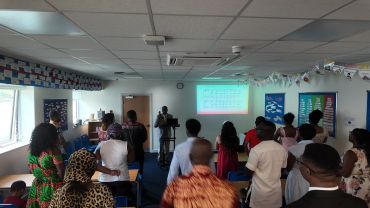 A group of people standing in a classroom with a man speaking at a podium and music notes projected on the wall.