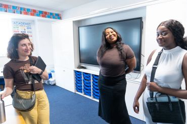 Three women standing and chatting in a bright room with a large screen.