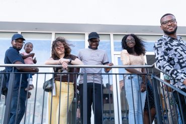 Diverse group of six people smiling and standing on a balcony railing outdoors.