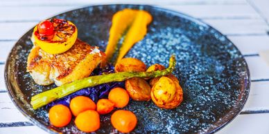 Gourmet fish fillet with grilled lemon, asparagus, and colorful vegetables on a black plate.