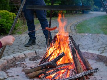 Sausage roasting over open campfire in backyard setting, people enjoying outdoor cooking with flames
