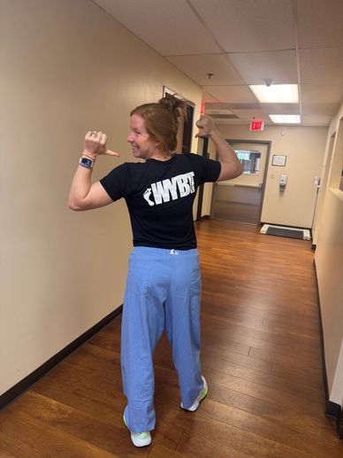 Woman in blue scrubs pointing at 'WYBT' on her shirt.