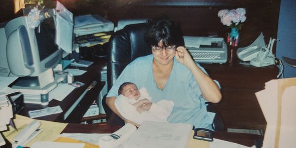 A woman holding a baby while sitting at a desk with office equipment.