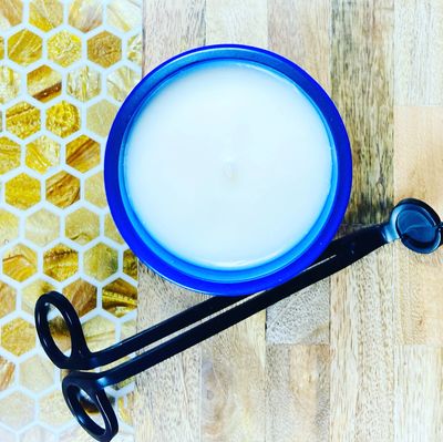 A white candle in a blue holder with honeycomb background and black wick trimmer.