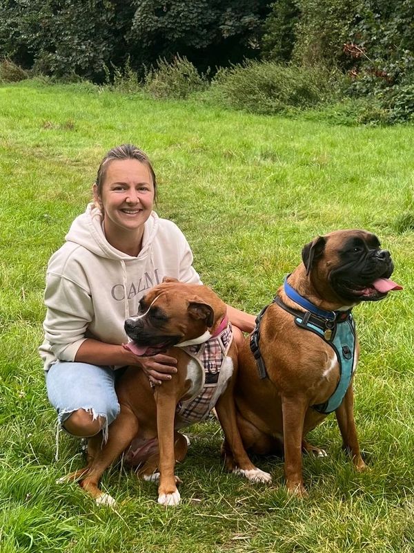 Smiling woman kneels with two brown dogs in a grassy field with trees in the background.