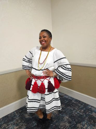 Woman in traditional white dress with black zigzag patterns and red tassels smiles confidently.